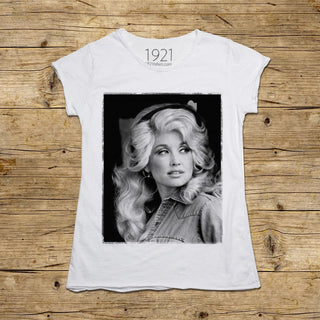 A white short-sleeve graphic T-shirt laid flat on a rustic wooden surface. The shirt features a large black-and-white portrait print of Dolly Parton centered on the front, with '1921' and 'dollyparton.